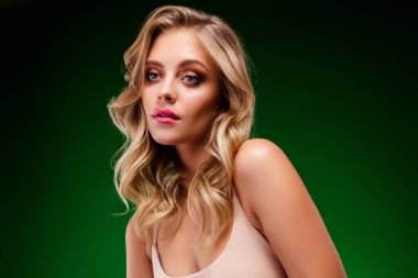 Güzel, sarı saçlı bir kadın şık giyinmiş, mükemmel bir cilde sahip, stüdyo ışıklandırmasında canlı yeşil bir arka plana poz vermiş, zarif ve modern güzelliği temsil eden bir şekilde tasvir ediliyor..