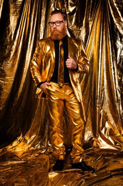 Kırmızı sakallı ve gözlüklü zarif bir beyefendi gösterişli altın bir takım elbise içinde duruyor. Bayram kutlaması için klasik bir cazibe yayıyor..
