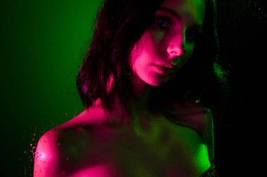 Yeşil ve pembe renkli dramatik ışıklandırma altında genç bir kadının yaratıcı neon portresi yüzeyinde su damlacıkları ve doku ve entrika ekleyerek sanatsal ve modern bir estetik tasvir ediyor..