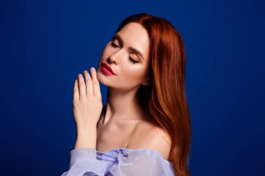 Mükemmel makyajlı, kusursuz ciltli, parlak mavi arka planda zarif duruşu olan kızıl saçlı genç bir kadının yakın çekim portresi.