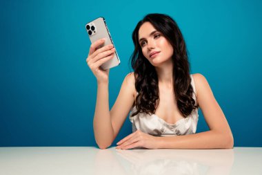 Zarif bir kadın akıllı telefon tutuyor, şık kıyafetler giyiyor, canlı mavi bir arka planda oturuyor, özgüven ve moda tasviri yapıyor..