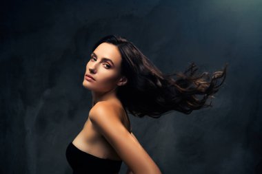 Koyu dokulu arka planda uzun dalgalı saçları olan, güzelliğini, zarafetini ve cazibesini ön plana çıkaran zarif bir kadın portresi. Moda, makyaj ve cilt kavramları için uygundur..