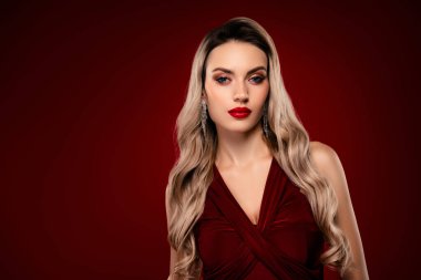 Zarif kırmızı elbiseli, sarı saçlı ve bordo bir arka planda poz veren alımlı genç manken.