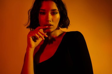 Kısa esmer saçlı, etkileyici pozlu, turuncu ve sarı renkli stüdyo ışıklarıyla yıkanmış şık bir kadının yakın çekimi..