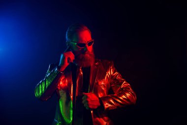 Olgun bir adam parlak bir takım elbise giyer ve güneş gözlüklerini parlak mavi ve kırmızı neon ışıklarla ayarlar. Koyu bir arka plana karşı zarafet ve karizma yayar..