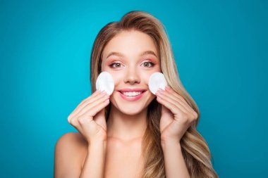 Uzun sarı saçlı, yanaklarında pamuk pedlerle çekici genç bir kadın, gösterişli cilt bakımı, güzellik rutini, ve canlı mavi bir arka planda zarif doğal cazibe..