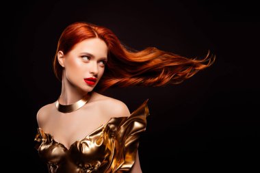 Zarif, kızıl saçlı, göz alıcı makyajlı altın rengi bir elbise giyen ve parlak siyah bir arka planda poz veren genç bir kadın..