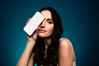 Yüzünün yanında boş ekranı olan bir akıllı telefonu tutan esmer kadın, mavi eğimli bir arkaplan üzerine kurulmuş. Yaratıcı projelerde teknoloji, güzellik ve moda temaları için mükemmel..