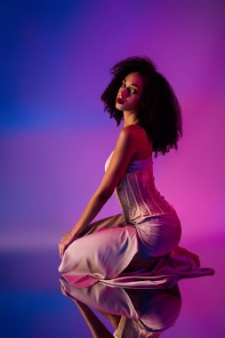 Büyüleyici bir portre, göz alıcı bir elbise içindeki genç, şık bir kadın, ışıltılı kıvrımlar ve canlı neon stüdyo ışıklandırması ile kendine güvenen poz.