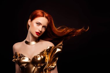 Kırmızı saçlı, altın sarısı, sanatsal bir elbise giyen, ışıl ışıl ve güzel, parlak ışık altında siyah, siyah bir arka plana karşı parıldayan genç bir model..