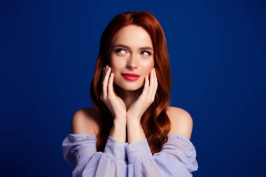 Genç kızıl saçlı bir kadın zarif bir şekilde mavi bir arkaplanda poz veriyor. Şık bir güzelliğin ve modern zarafetin simgesi. Parlak teni ve kendine güvenen mükemmel makyajı ve saç stili.