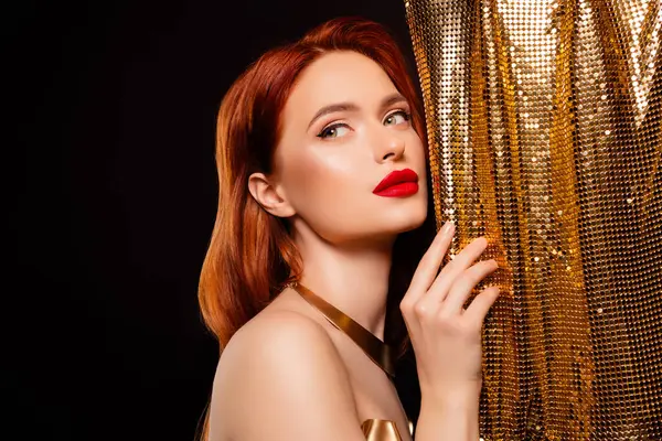Göz kamaştırıcı kızıl saçları ve göz kamaştırıcı altın rengi elbisesi olan büyüleyici genç bir kadın zarif, siyah bir arka plana karşı duruyor. Zarafet ve cazibe saçıyor..