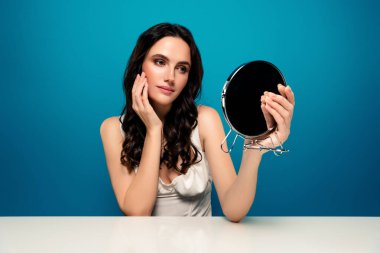 Elinde ayna tutan esmer bir kadın, eğimli mavi arka planda otururken tenini inceliyor. Zarif duruşu ve doğal güzelliği dingin ve şık bir estetiği vurguluyor..