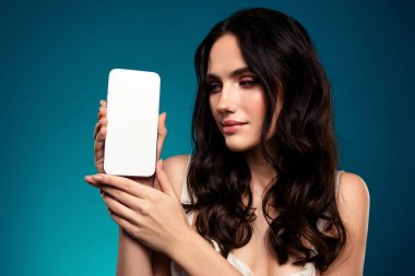 Güzel, genç, esmer bir kadın, beyaz bir akıllı telefonu mavi bir arka plana karşı sergiliyor, modern teknoloji ve stili vurguluyor..