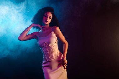 Yumuşak neon ışıkta zarif poz veren zarif genç bir kadın. Şık kıyafeti, kendine güveni, cazibesi ve şatafatlı zarafeti canlı ve karamsar ortamda..