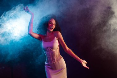 Zarif kıyafetler giyen çekici genç bir kadın renkli neon ışıklar ve atmosferik dumanlar arasında neşe içinde dans ediyor. Sanatsal bir stüdyo ortamında, kendine güven ve canlılık yayıyor..