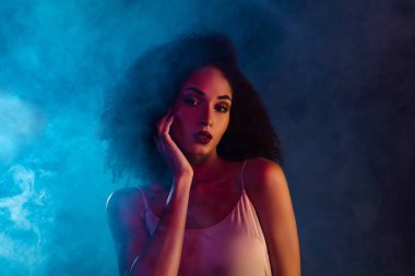 Gösterişli bakışları ve kıvırcık saçları olan gizemli dumanlı atmosferli neon ışıkları altında kendine güveni ve güzelliği olan zarif genç bir kadın..