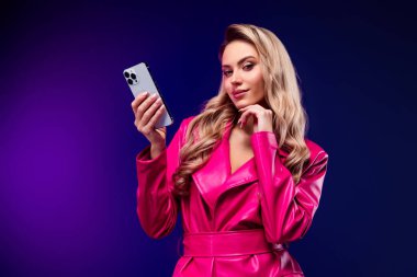 Şık, sarı saçlı bir kadın pembe bir trençkot giyiyor, elinde parlak mor bir arka plan, ışıldayan bir güzellik, kendine güven ve modern yaşam tarzından ilham alan bir akıllı telefon tutuyor..