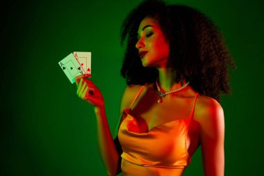 Şatafatlı moda kıyafetleriyle süslenmiş kendine güvenen genç bir kadın kartlar tutar, canlı yeşil bir arka plana karşı zarafet ve cazibe yayar. Cazibe, zarafet ve kumarhane tarzı için mükemmel bir tema..
