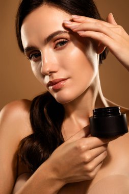 Siyah bir kavanozda doğal bej renkli pastel arka planı olan cilt ürününü sunan güzel bir genç kadının portresi..