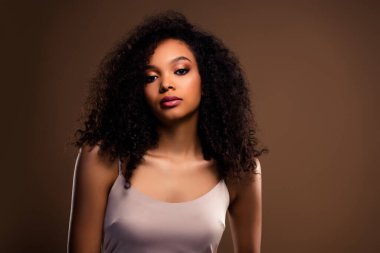 Kıvırcık siyah saçlı, çekici bir genç kadın. Kahverengi arka planda, minimal bir elbiseyle poz veriyor. Özgüveni ve tarzı inceliği ve zarafeti resmediyor..