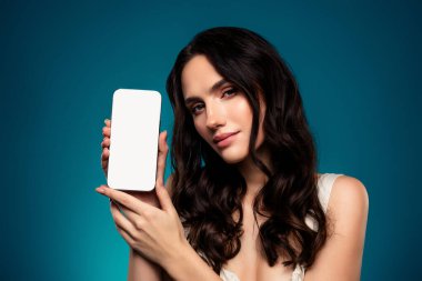 Güzel esmer bir kadın, şık bir arkaplan karşısında boş bir ekranla akıllı telefonuyla şık görünümünü, şık makyajını ve çekici saç stilini sergiliyor..