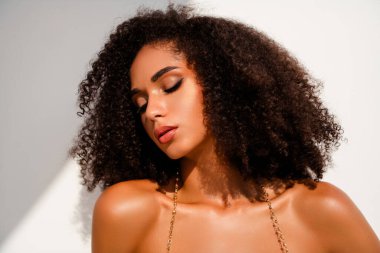 Şaşaalı kıvırcık saçlı ve şık makyajlı zarif genç bir kadın beyaz, parlak bir arka plana karşı zarifçe poz veriyor..