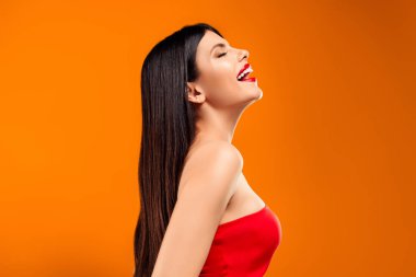 Genç bir kadın kırmızı bir bluzla poz veriyor, mutluluk saçıyor ve parlak turuncu bir arka plana karşı güzel saç stili ve makyajını sergiliyor.