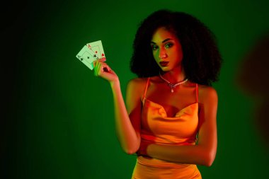 Çekici bir kokteyl elbisesi giymiş kendine güvenen genç bir kadın, elinde parlak yeşil bir arka plana karşı duran kart tutarken şık bir ifade sergiliyor..