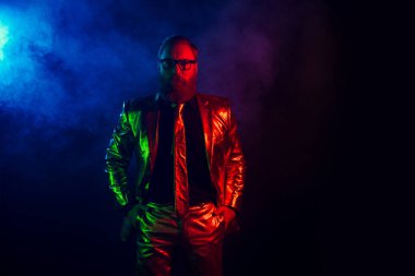 Parlak neon takım elbiseli zarif, karizmatik bir adam karanlık bir arkaplana karşı poz veriyor, eski tarz ve kendine güvenen bir tavır sergiliyor, gece kulübü veya parti temaları için mükemmel..