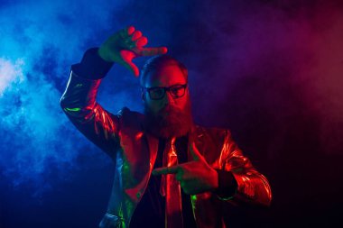 Sakallı ve gözlüklü olgun bir adam esmer, dumanlı bir arka plana karşı parlak bir takım elbiseyle poz verir..