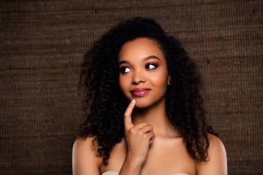 Çekici, kıvırcık saçlı ve zarif makyajlı genç bir kadın kahverengi bir arkaplanda, kavramsal bir moda imajıyla güzelliğini ve tarzını sergiliyor..