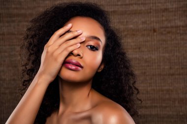 Zarafet ve güzellik sergileyen genç bir kadın bir gözünü şık bir şekilde kapatıyor. Makyajı ve kıvırcık saçları doğal kahverengi bir arka plana karşı duruşunu arttırıyor..