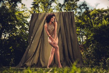 Leopar desenli kadın zarif bir poz veriyor, yemyeşil kumaşlara karşı, yemyeşil bir bahçede, ılık güneş ışığı altında..