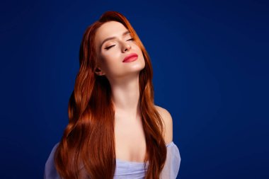 Genç, kızıl saçlı, parlak saçlı ve kapalı gözlü bir kadın zarif bir şekilde poz veriyor, bir güzellik hissi yayıyor ve canlı, mavi bir arka plana kurulup zarif ve zarif özelliklerini vurguluyor..