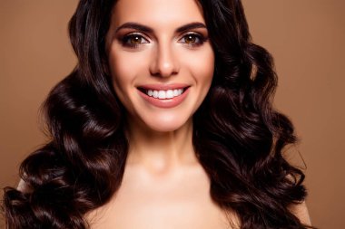 Işıl ışıl gülümseyen esmer kadın, göz kamaştırıcı makyajı ve bej bir arka plana poz veren şehvetli kıvırcık saçları, coşkulu bir güzelliği ve güveni var..