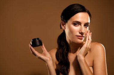 Yüzüne dokunurken elinde bir kavanoz tutan esmer saçlı genç bir kadın, bej bir arka plan üzerine kurulmuş, güzellik ve özen gösteren.