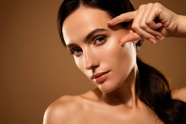 Yüzüne dokunan genç bir kadının yakın plan çekimi, bej bir arka planda doğal güzelliğini ve cildini vurgulaması, zarafet, güven ve refah hissi yayması..