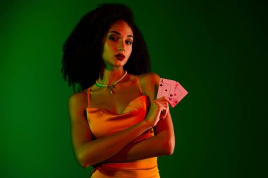 Göz kamaştırıcı, şık giyinmiş genç bir kadın elinde iskambil kâğıdı, çarpıcı yeşil bir arka plan, ihtişamlı, sofistike ve stil temaları ile kendinden emin bir şekilde poz veriyor..