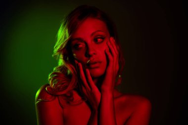 Genç bir kadın koyu bir arkaplanda neon kırmızı ve yeşil ışıkla poz veriyor, güzelliğini ve şehvetini çıplak omuzları ve sarı saçlarıyla vurguluyor..