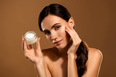 Güzel, genç, esmer bir kadın sıcak pastel arka planda krema kavanozu tutuyor, zarafet ve zarafetle güzellik bakımını sergiliyor..