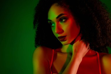 Genç bir kadın göz kamaştırıcı bir poz veriyor canlı yeşil ve kırmızı ışıklı bir ortamda, şık tarzını ve zarif varlığını vurguluyor..