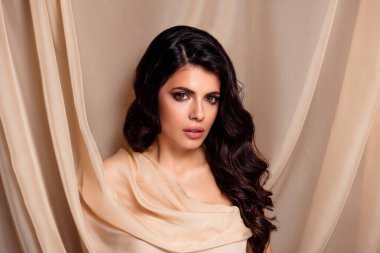 Güzel esmer kadın, doğal makyajlı, açık renk kumaş giyiyor, çekici bir saç stiliyle poz veriyor ve bej bir arka plana karşı büyüleyici bir ifade sergiliyor, kendine güveni ve kadınsılığı var..