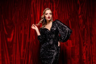 Siyah pullu bir elbise içinde şık bir genç kadın zengin kırmızı kadife bir perdenin önünde duruyor, büyüleyici bir duruşu ve kendine güveni var..