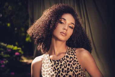 Kıvırcık saçlı ve leopar desenli elbiseli genç bir kadın bahçede duruyor, yumuşak güneş ışığıyla yıkanmış. Dekor onun güzelliğini ve doğayla bağlantısını vurguluyor..