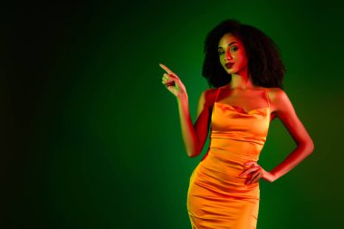 Kendine güvenen turuncu elbiseli bir kadın canlı yeşil bir arka plana karşı duruyor, zarafet, moda ve modern tarzı sergiliyor..