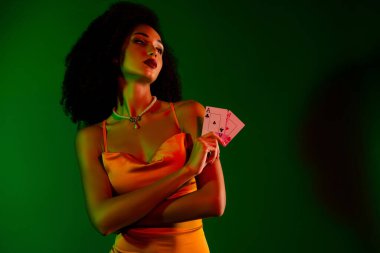 Elinde poker kartları olan şık bir kokteyl elbisesi giymiş şık bir kadın. Lüks, moda ve cazibe temalarını canlı yeşil eğimli bir zemine karşı yakalar.