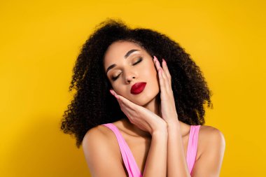 Kıvırcık saçlı ve parlak kırmızı rujlu genç bir kadın zarif bir şekilde sarı arka plana karşı poz verir, kendine güveni ve güzelliğini şık ve büyüleyici bir şekilde ifade eder..