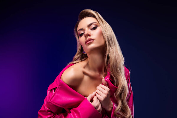 Elegant young woman with long blonde hair wearing a pink coat, posing against a vibrant purple and violet background, showcasing beauty and style in a sensual and confident manner.