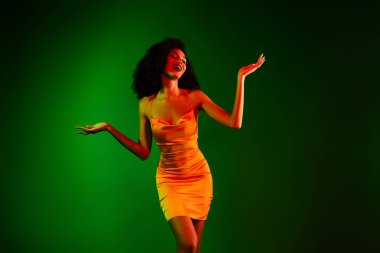 Turuncu bir gece elbisesi giymiş genç bir kadın enerjik bir şekilde poz veriyor. Parlak yeşil bir arka planı var. Lüks ve cazibeyi resmediyor..